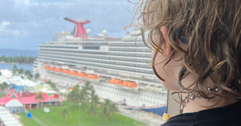 A young person with curly hair and a necklace is looking out a window at a Carnival cruise ship docked in port.