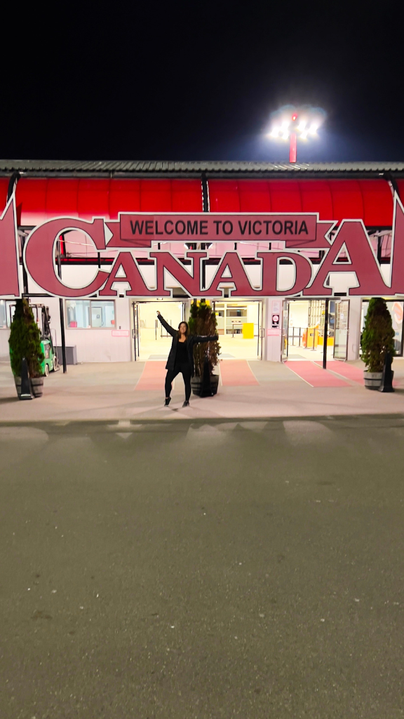 A person standing under a large "Welcome to Victoria Canada" sign at night. The sign is illuminated, creating a welcoming atmosphere as the person waves enthusiastically.