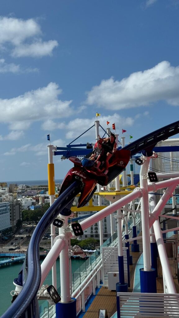 Two people riding an exciting roller coaster on a cruise ship with a scenic city view in the background.